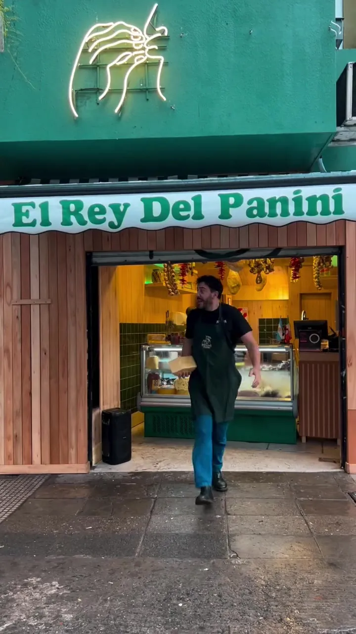 La fachada del local: toldo verde con letras crema que dice El Rey Del Panini, revestimiento de madera y el mostrador iluminado